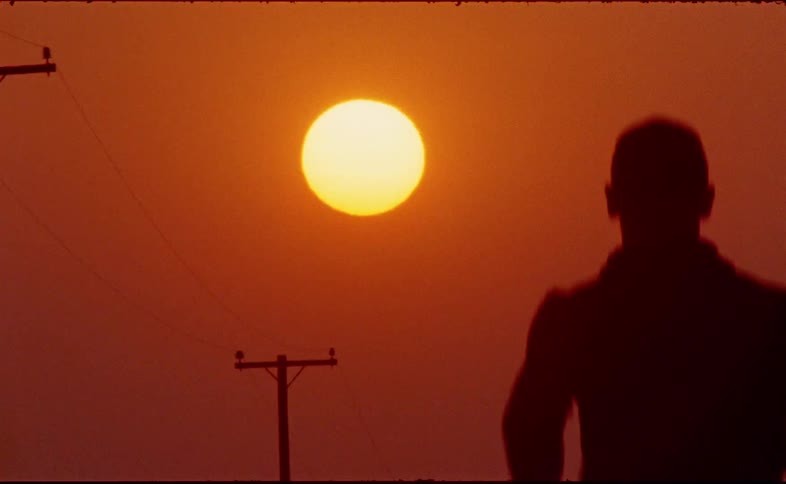 A person in silhouette walks away from the camera toward the sun in a vibrant orange sky