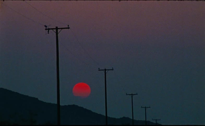 A brilliant red sun sets behind silhouetted hills and a line of telephone poles in a grainy time-lapse