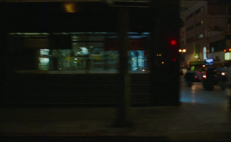 A grainy, drive-by view of a quiet downtown Los Angeles street at dusk, with traffic, pedestrians, and glowing neon signs