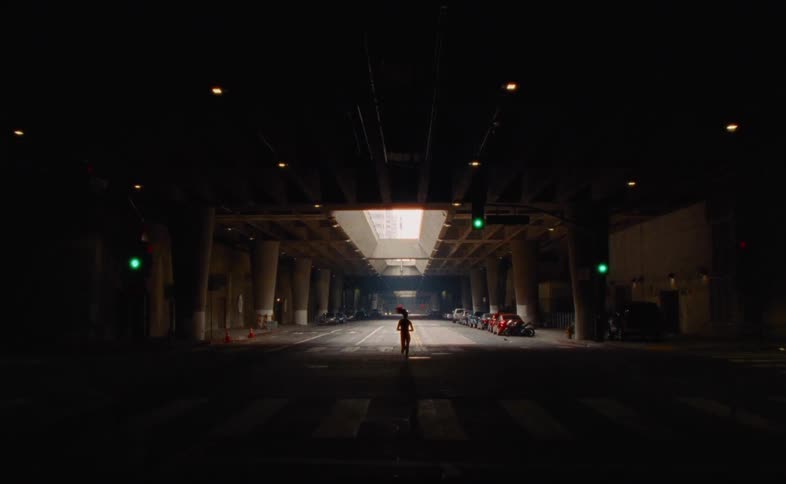 A silhouetted woman runs in slow motion down the center of an empty city street under a dark overpass, moving towards the light