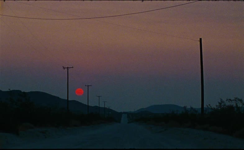 A stationary view of a long, straight desert road where a deep red sun sets behind distant mountains