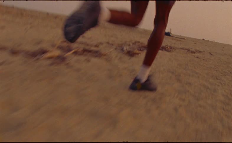 A low-angle tracking shot follows the legs and feet of a person running on a sandy beach during a golden sunset