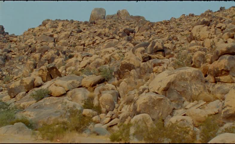 Slow pan reveals a vast and rugged desert landscape with a hillside covered in large, weathered boulders