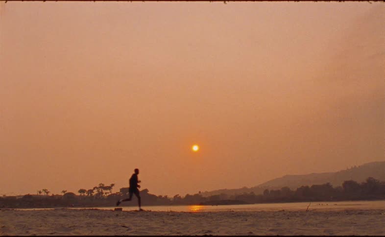 A silhouetted runner jogs along the sandy shore of a river under the warm glow of the setting sun