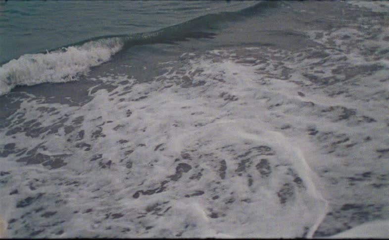 Vintage Super 8 film captures gentle ocean waves washing onto the sandy shore in slow motion
