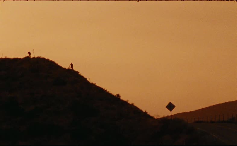 Two silhouetted figures meet on a dusty hillside against a hazy, orange-toned sky