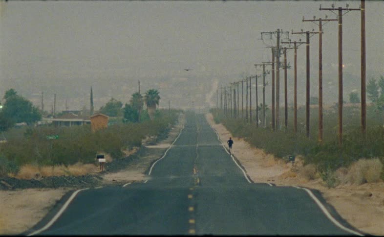A solitary figure walks down a long, empty desert road lined with telephone poles on a hazy day