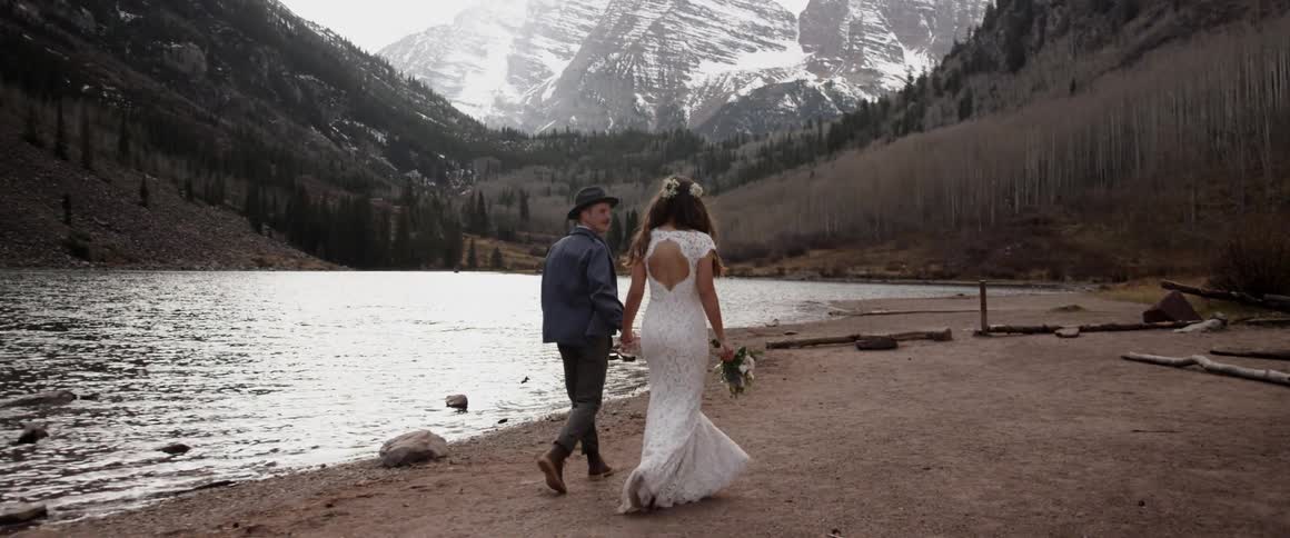 A bride and groom walk hand in hand by a mountain lake
