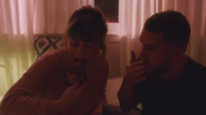 A man and woman sit in bed and eat some food
