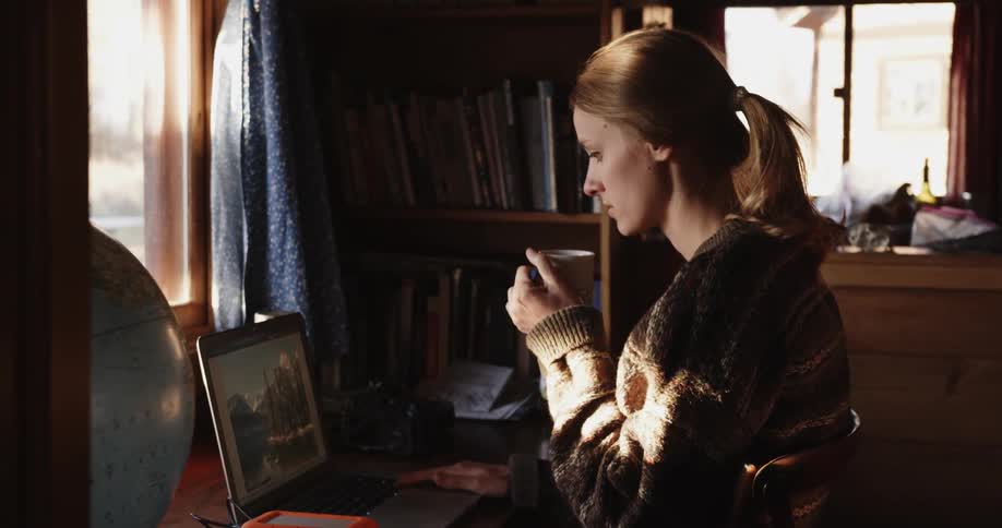 A woman sits by her laptop sipping from a mag