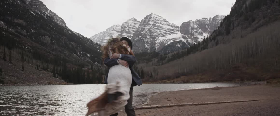 A groom spins his bride around near a mountain lake