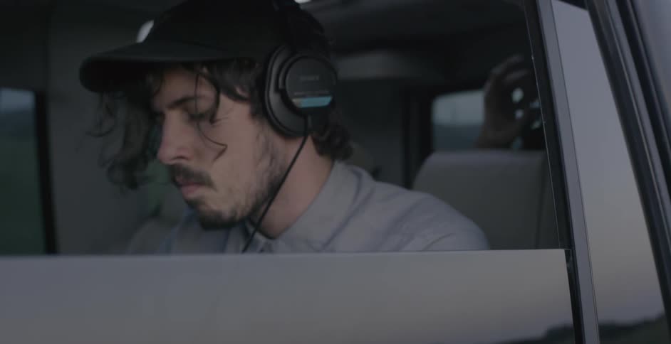 A man in a car looking around as he rides on the back and takes his headphones off and puts them on