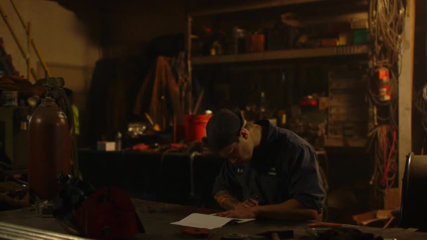 A man sits at a desk in a garage drawing on a pad of paper