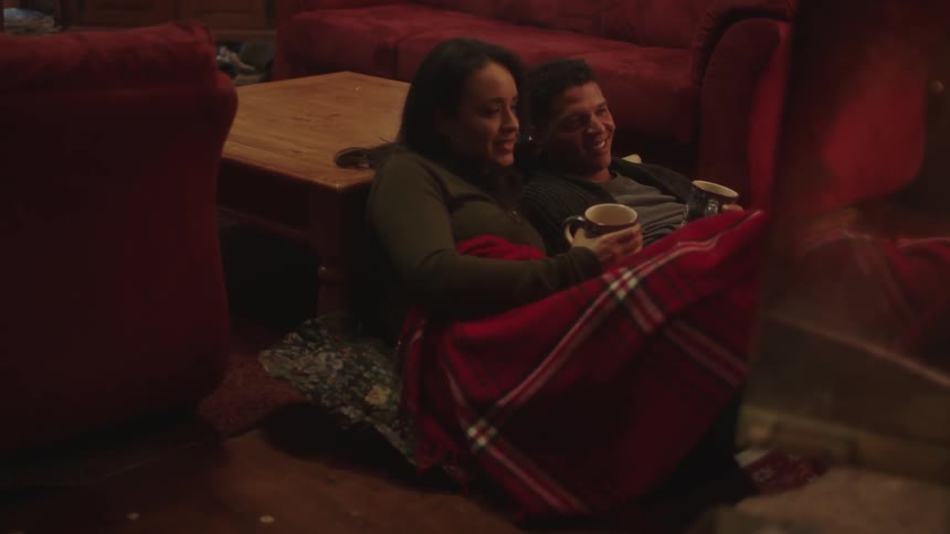 A man and woman share a blanket while drinking from mugs on a living room floor