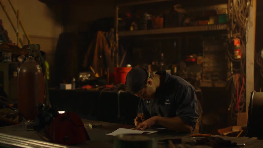 A man draws in a notebook while sitting at a workbench in a shop