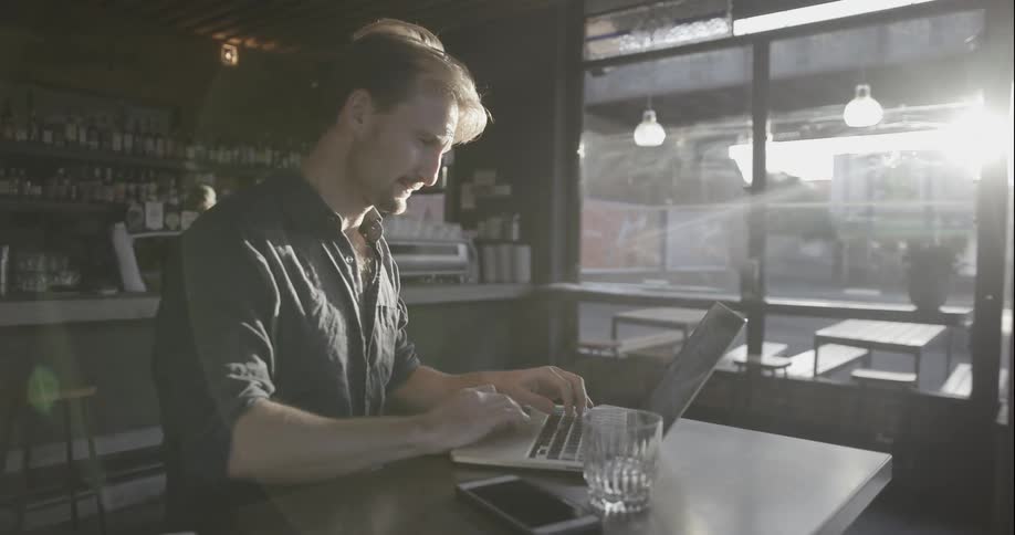 A man takes a drink looking at his laptop in a cafe