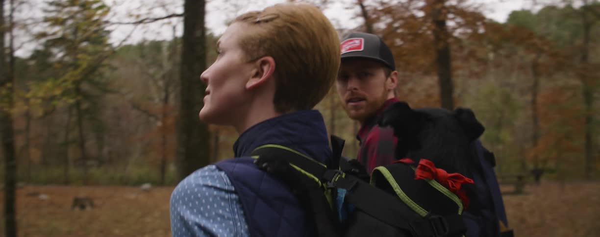 Man and woman walk together in the woods while woman holds dog in a pack