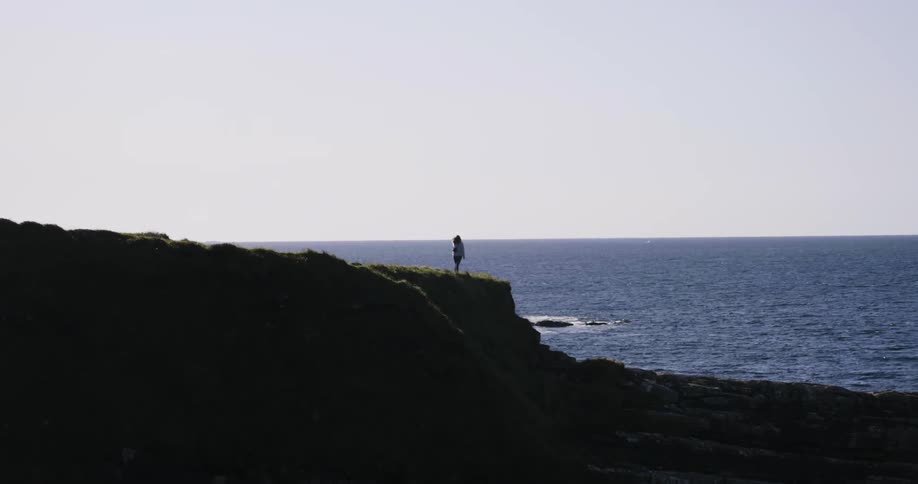 A person stands on the edge of a cliff by the water