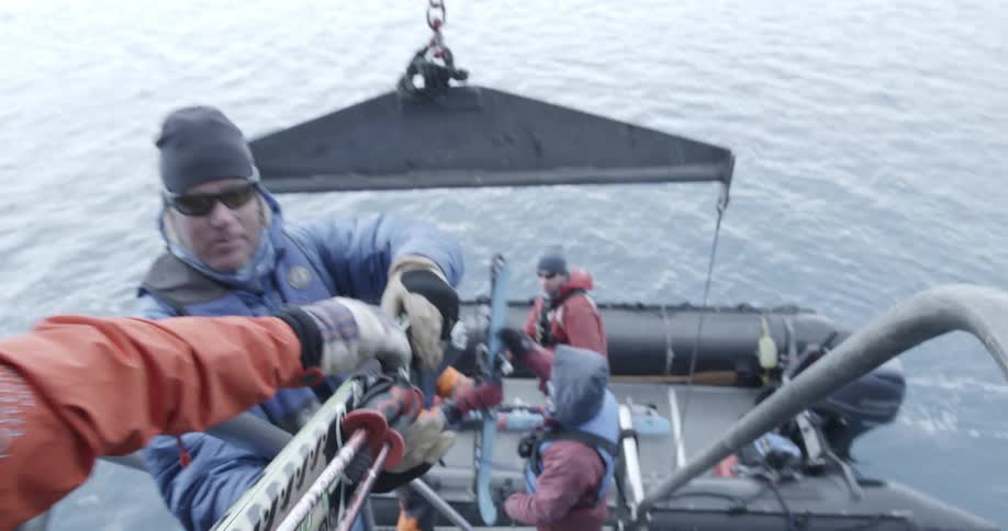 A man hands skis from a ship to a man in a zodiac