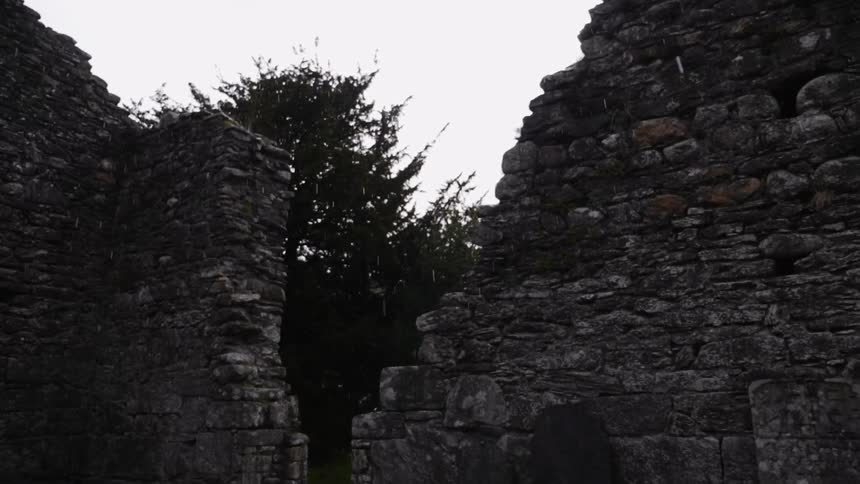 Snow falls over the crumbling ruins of a stone building during the day