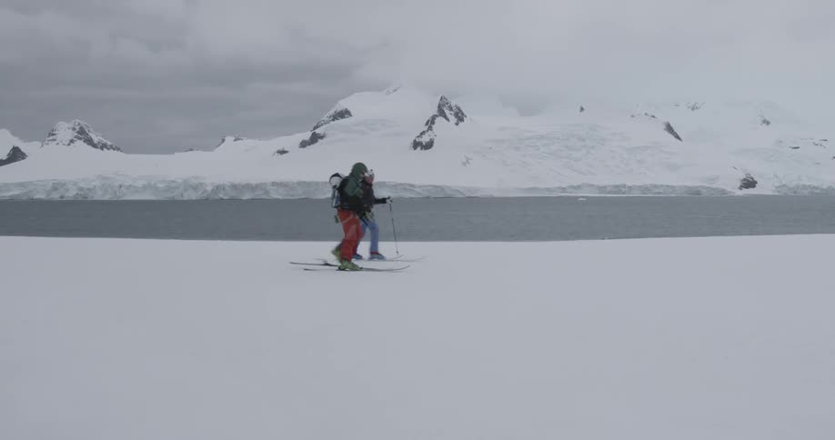 Two people ski walk across a flat snowy area (tilt up)