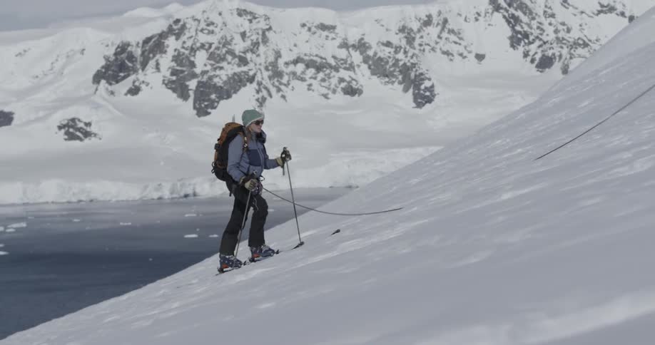 A woman hikes up a snowy mountainside with skis (wide)
