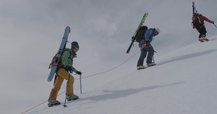 People negotiating a snowy hill climb tethered together and using walking poles, ice axe, and crampons