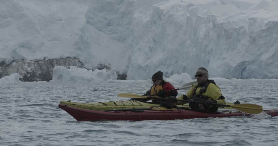Two men kayak through the ocean with ice chunks and glaciers in the background