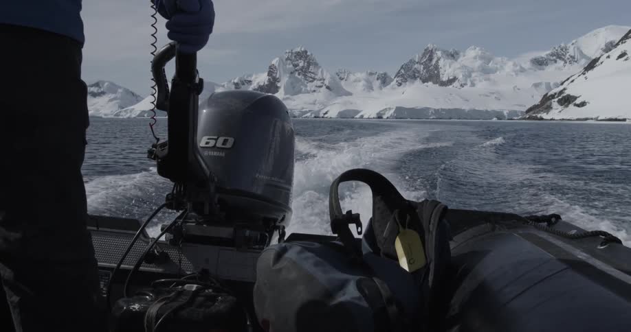 A person steers an outboard boat through icy water surrounded by snowy mountains
