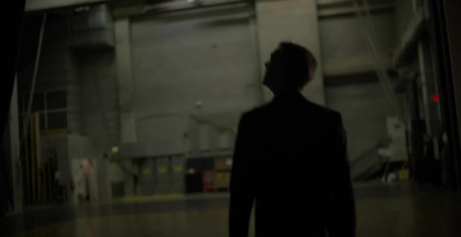 A man stands in a dark warehouse before walking forward