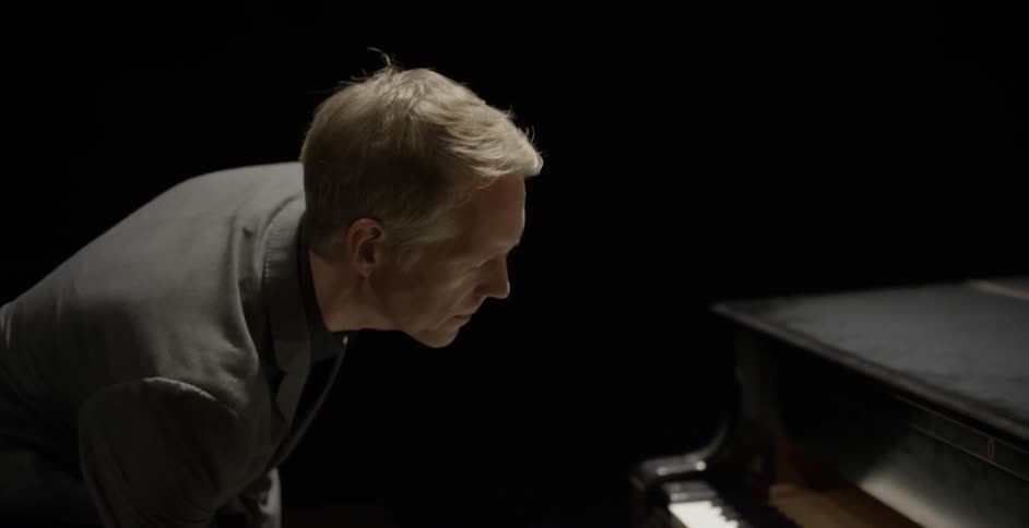 A man walks onto an auditorium stage and sits down at a piano