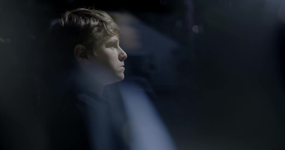 Boy stands looking out at night while a light shines on him