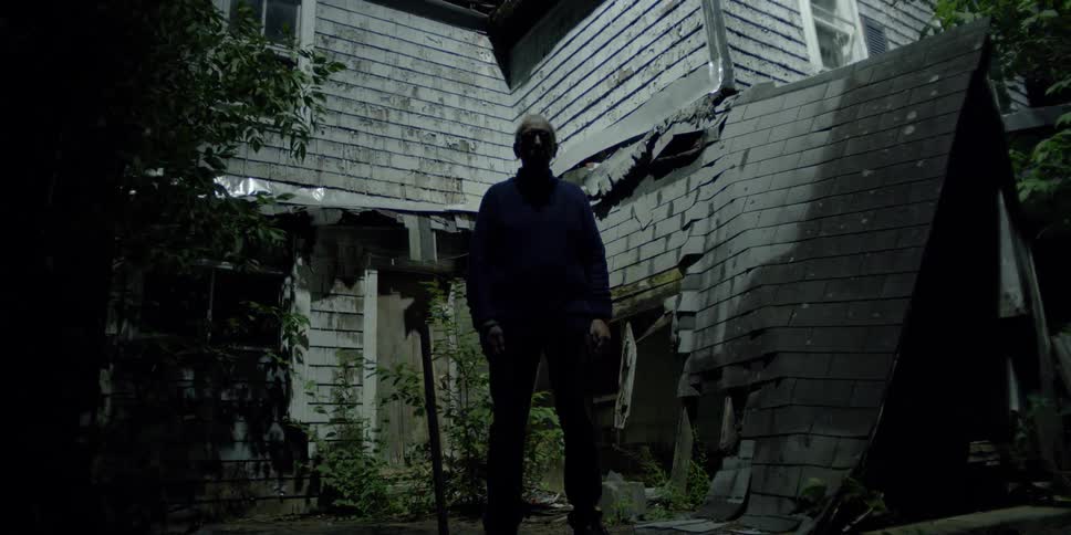 An old man stands still outside a dilapidated old building at night