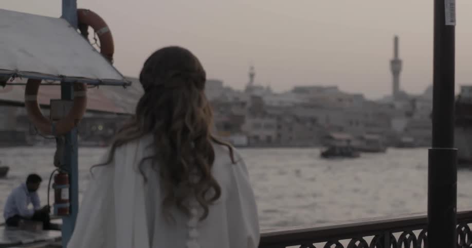 A woman stands watching boats on a waterway in a foreign city at dusk