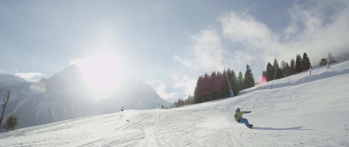 A snowboarder rides down a mountain in a ski resort during the day