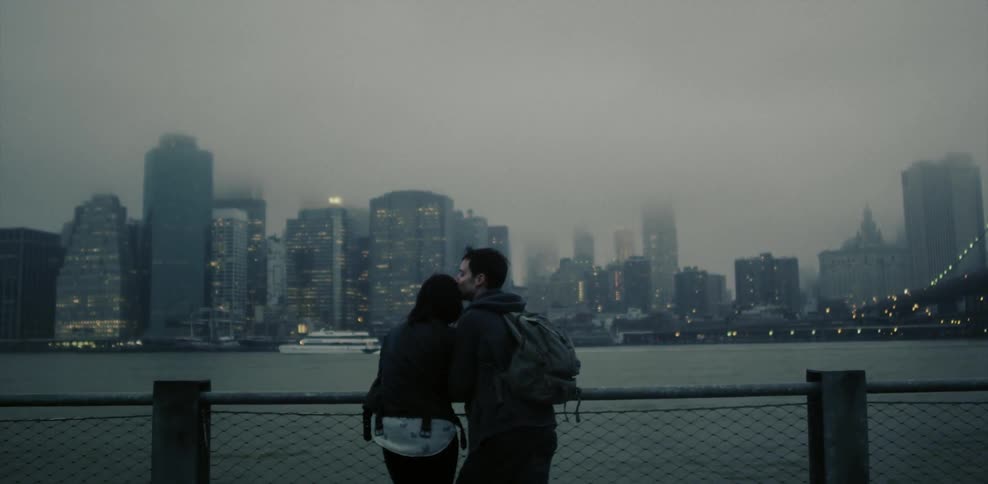 Couple standing against a rail across a river from a big city on a cloudy day