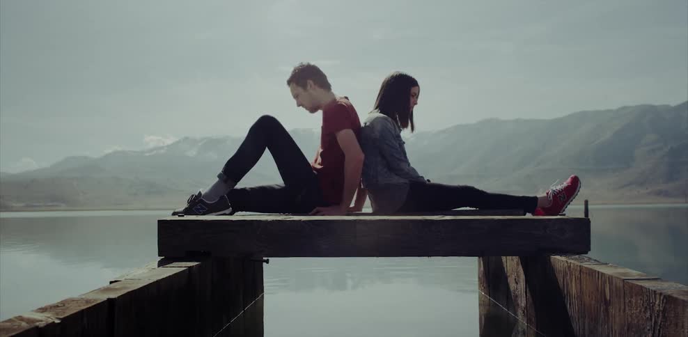 A man and woman are sitting back to back on a dock at a lake during the day