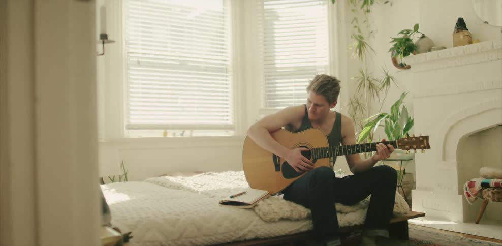 A guitarist holds a guitar and writes on paper on a bed