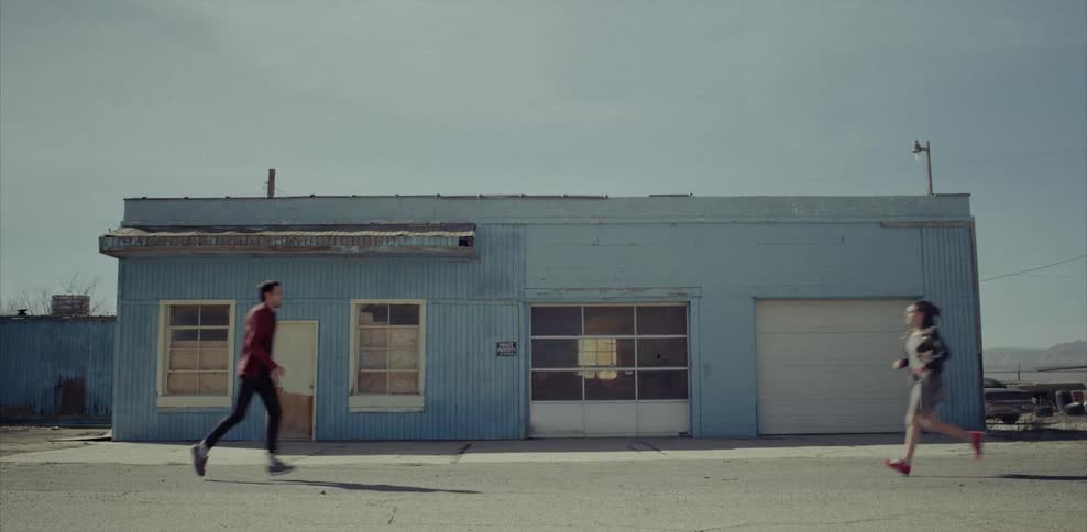 A man and a woman run up to each other outside a rundown garage in the daylight