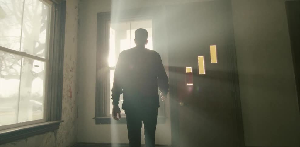 Man walking through a tattered house and goes to a window with beams of light shining in from a sunset