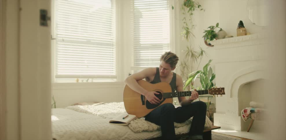 A man sitting on a bed with a guitar and writing on paper