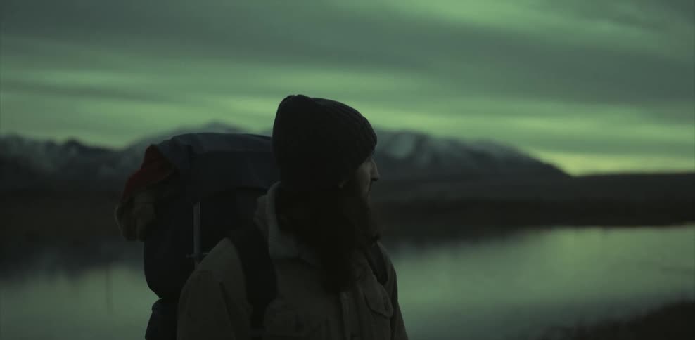 A bearded man wearing a backpack near a lake turns around