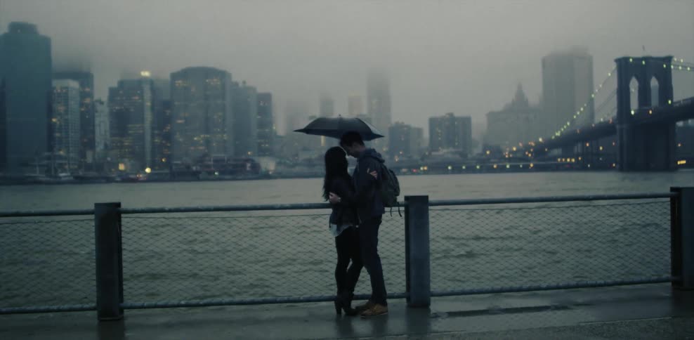 Man and woman hug under an umbrella next to a river and city