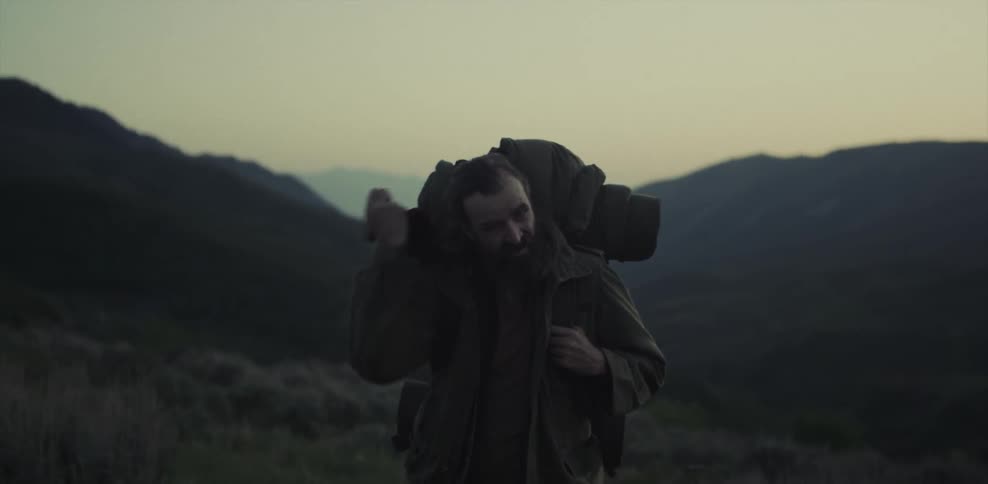 Man wearing a backpack stands on hilly terrain wiping his head