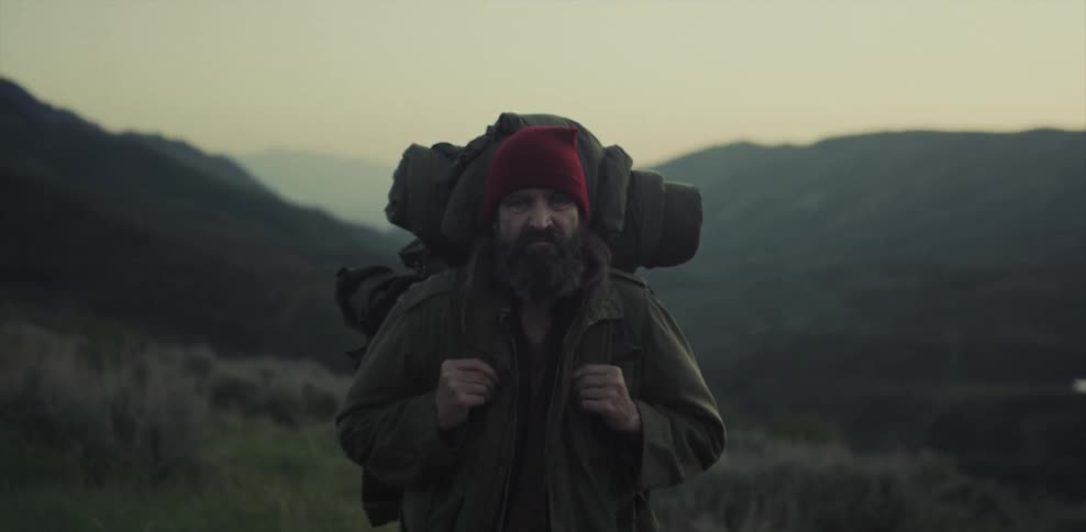 Frowning elderly man wearing a backpack stands still outside