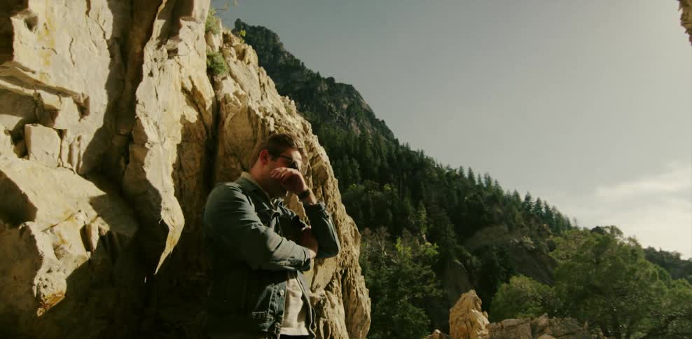 A man leans against the rock ledge and looks up at the sun
