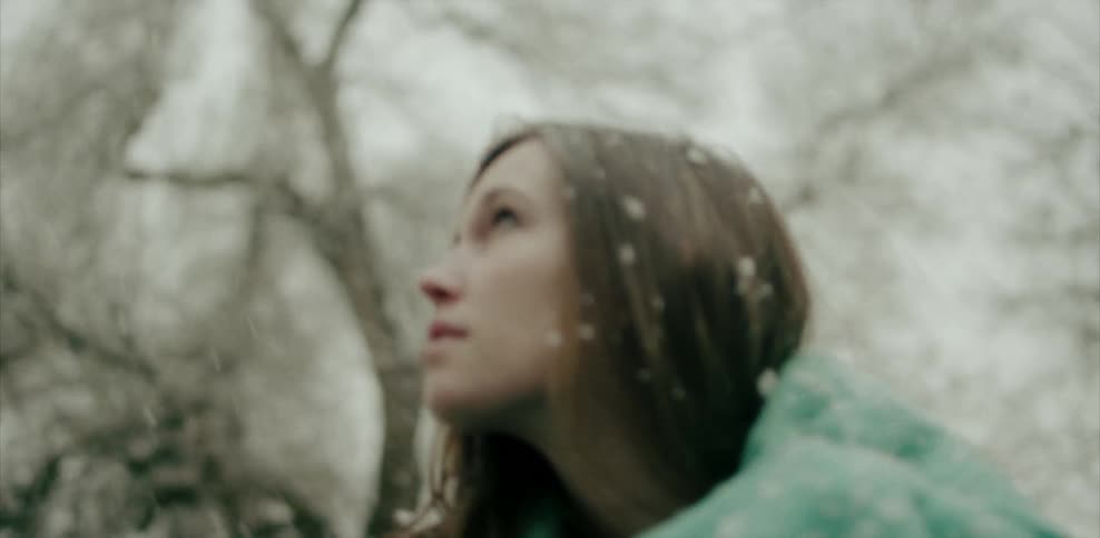 A woman covered in a blanket in the yard looking up at the snowy trees