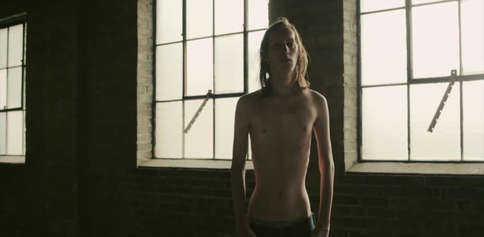 A thin man wearing jeans stands in front of a window