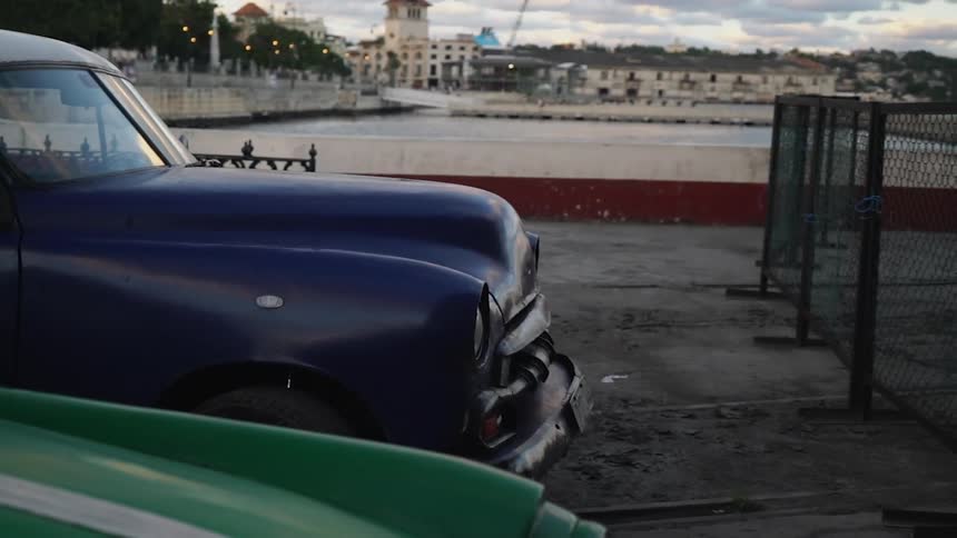 Two 1950s american cars sit parked near a harbor on a cloudy day