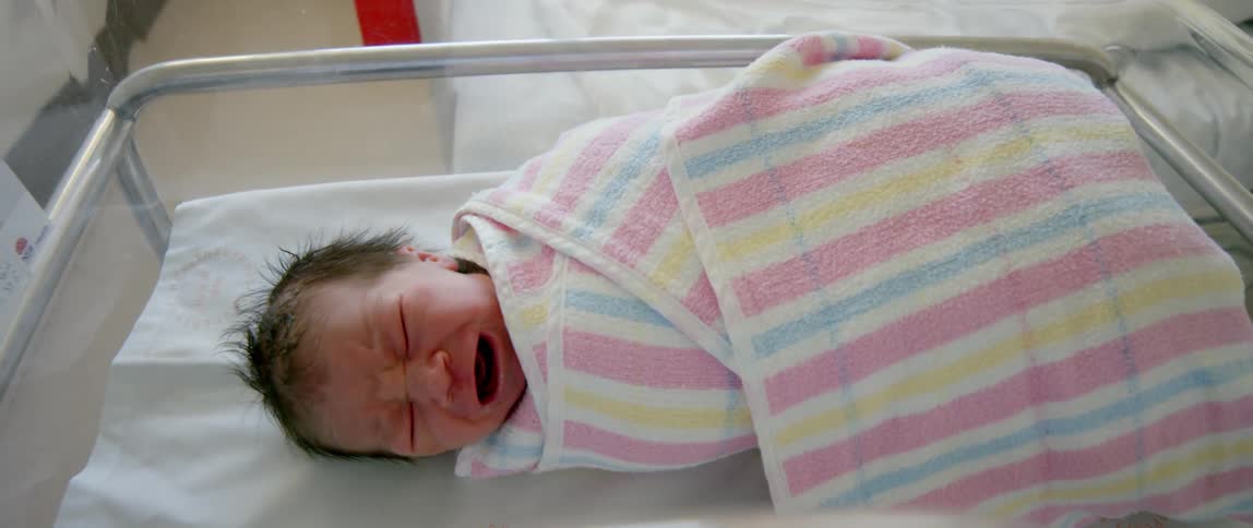 A swaddled newborn baby lies crying in its hospital crib